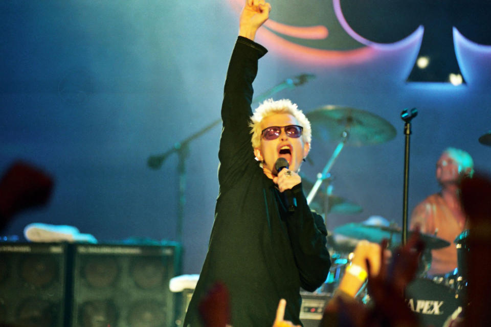 Billy Idol on stage singing into a microphone with his fist held high in the air.