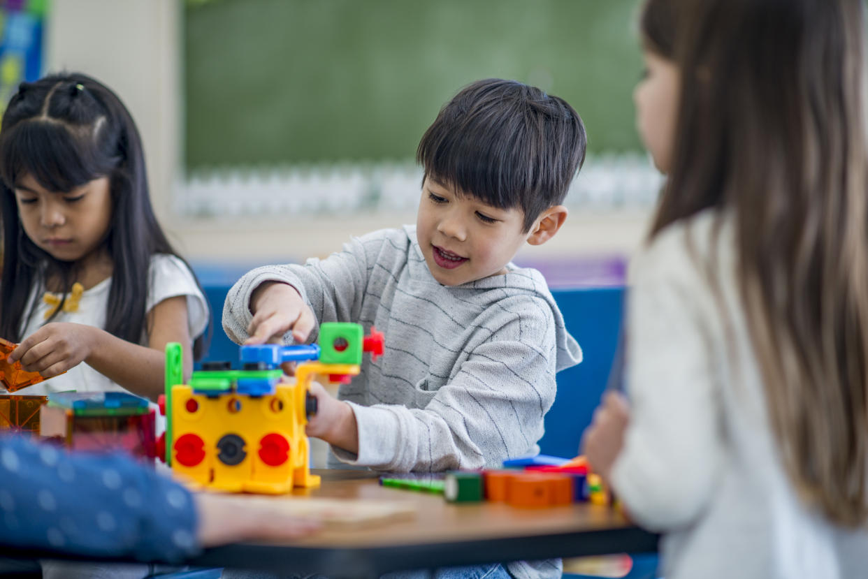 A group of preschool students are indoors in their classroom. They  are sitting with their teacher and playing with building blocks.