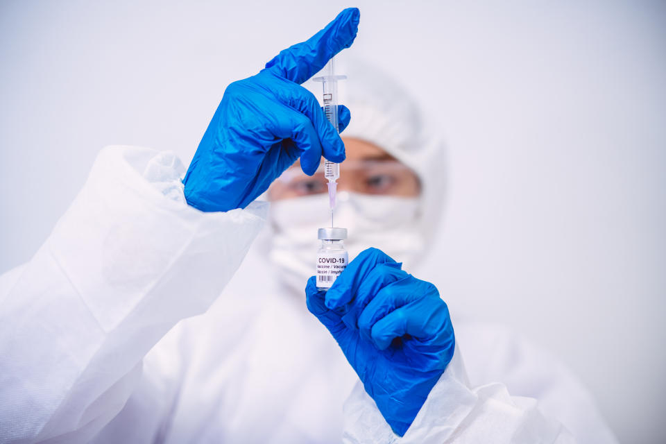 Doctor in protective gloves & workwear filling injection syringe with COVID-19 vaccine. 