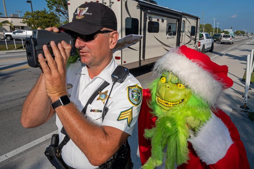 Lou Caputo, right, costumed as the Grinch, watches Sgt. Greg Korzan, left, as he uses a laser speed detector to check speeds of motorists traveling through a school zone on the Florida Keys Overseas Highway Tuesday, Dec. 13, 2022, in Marathon, Fla