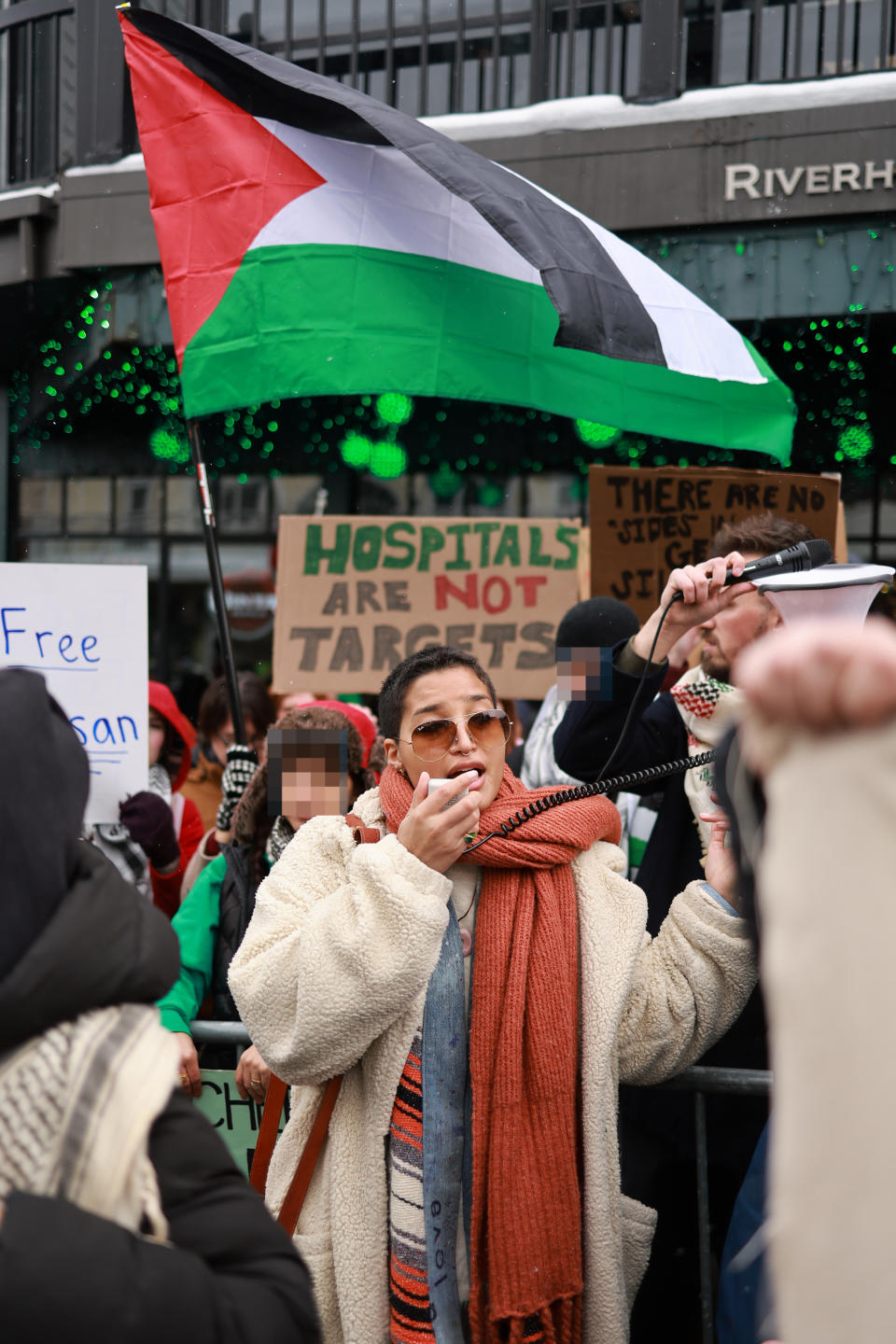 Protestors holding signs and Palestinian flags, with one woman in the front using a megaphone. Signs read, "Free" and "Hospitals Are Not Targets."