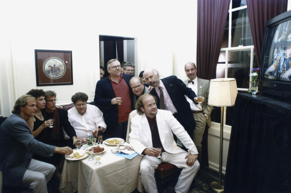 The cast and crew of <i>Cheers</i> gathers to watch the series finale. Pictured: (l-r) Woody Harrelson (Woody Boyd), Rhea Perlman (Carla LeBec), Ted Danson (Sam Malone), George Wendt (Norm Peterson), John Ratzenberger (Cliff Clavin), Paul Willson (Paul Krapence), Kelsey Grammer (Dr. Frasier Crane), and director/creator James Burrows (Photo: NBCU Photo Bank)