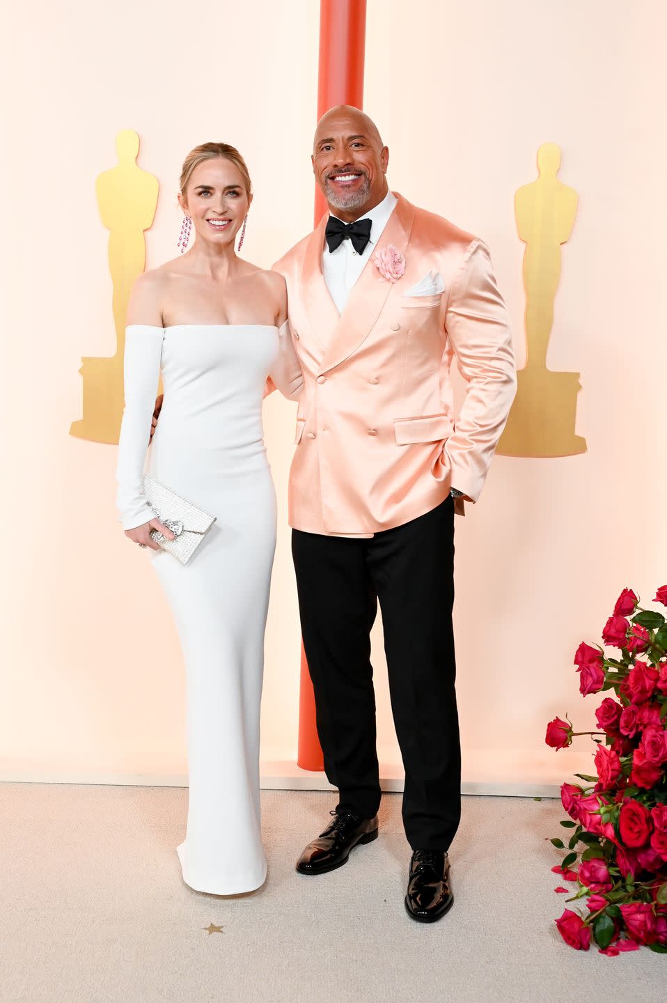 emily blunt in a strapless long white dress links arms with dwayne johnson in a black bowtie and trousers with peach satin tuxedo jacket