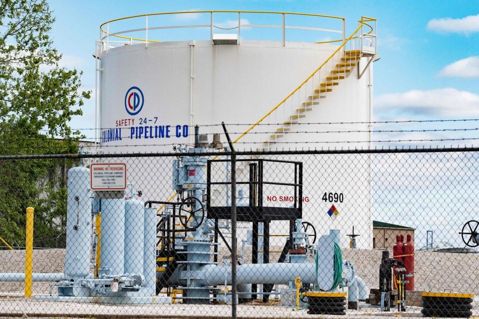 A chain-link fence topped with barbed wire in the foreground, large pipes and valves in front of a large white storage tank labeled Colonial Pipeline Co