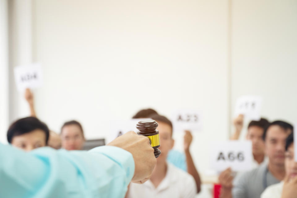 Close up auctioneer hand, holding gavel, wooden hammer, and blur group of people in auction room, one man raising hand up for bidding. Product or project auction market concept background (Close up auctioneer hand, holding gavel, wooden hammer, and bl