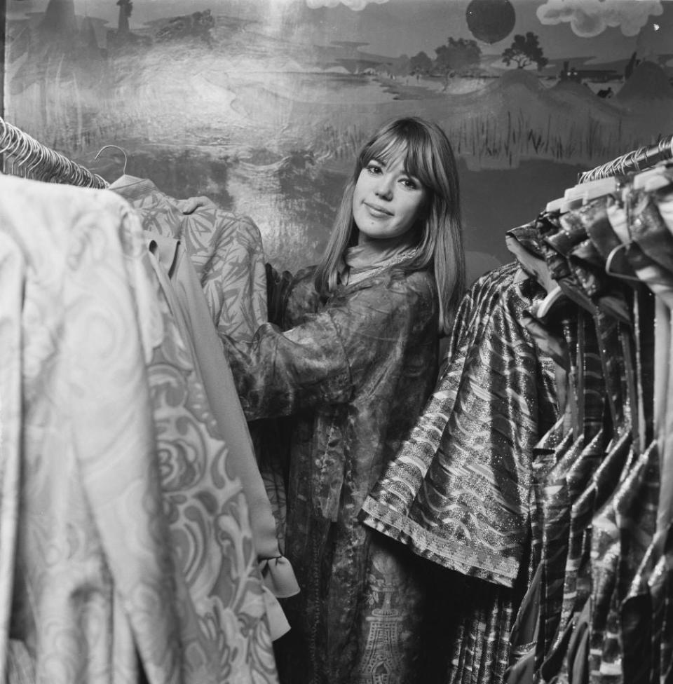 Jean Shrimpton looks through a rack of patterned clothing, set against an artistic backdrop