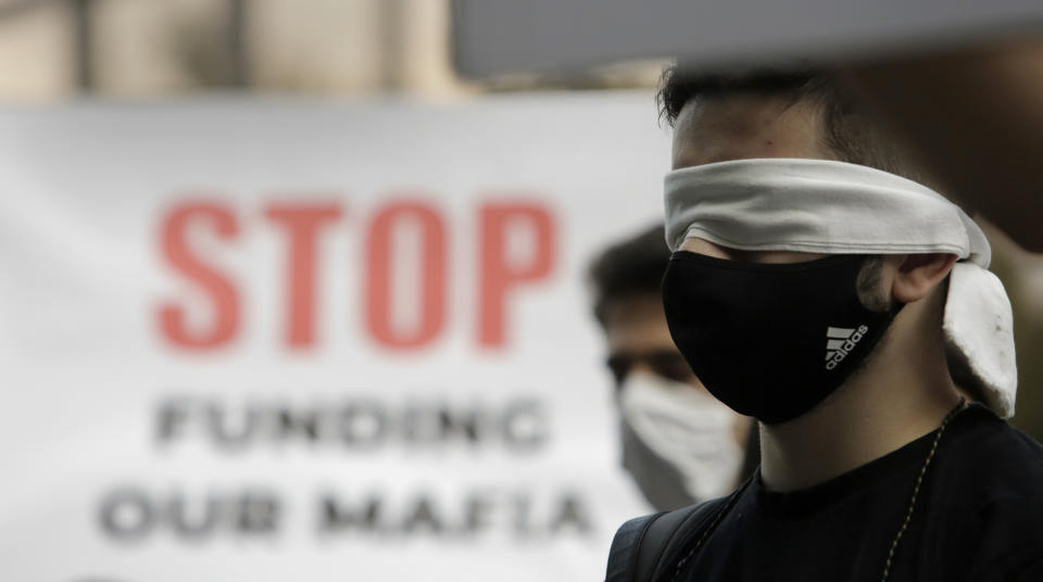 Protesters keep their eyes closed in front of a poster reading "Stop funding our mafia" in front of the German Embassy in Sofia, Bulgaria, Wednesday, Aug. 12, 2020. Several hundred anti-government protesters gathered in front of Germany’s embassy in Sofia, calling on Berlin and Brussels to “open their eyes” to widespread corruption in Bulgaria.The mostly young protesters accuse those in power of links to the mafia, refusing to fight corruption and suppressing freedom of speech. (AP Photo/Valentina Petrova)