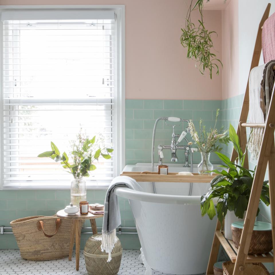 Pink and blue tiled bathroom with white bath, decorative storage baskets and house plants with big window and shutters