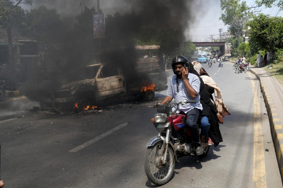 Una moto con varias personas a bordo circula junto a una camioneta incendiada por partidarios del ex primer ministro paquistaní Imran Khan, en Lahore, Pakistán, el 11 de mayo de 2023. (AP Foto/K.M. Chaudary)