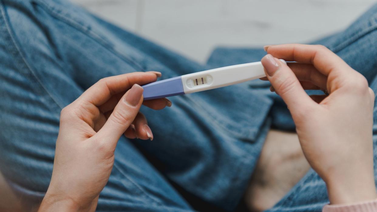  Young woman sitting on the bed and holding a positive pregnancy test. 