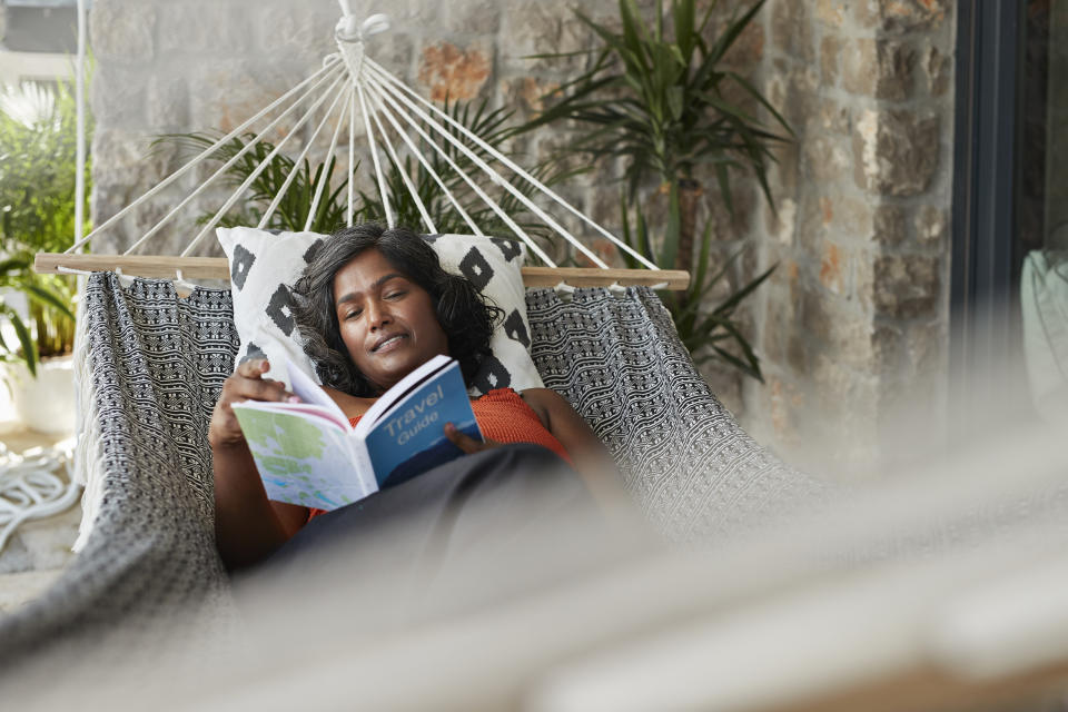 A woman is relaxing in a hammock, reading a book titled "Travel." The setting is a cozy patio with some plants in the background