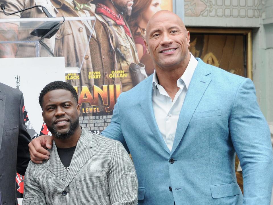 Kevin short in a grey suit and Dwayne much taller with his hand on Kevin's shoulder wearing a light blue suit.