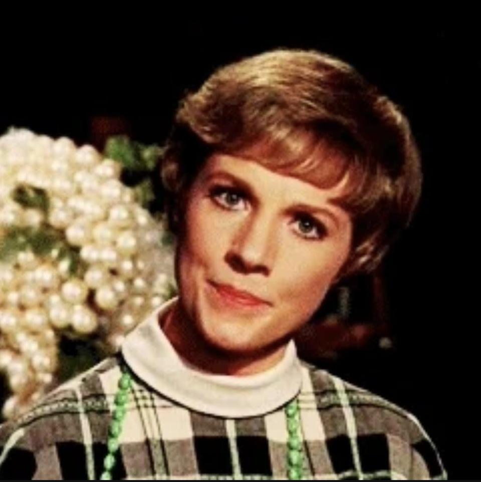 Julie Andrews wearing a checkered sweater and a necklace, standing in front of a floral background