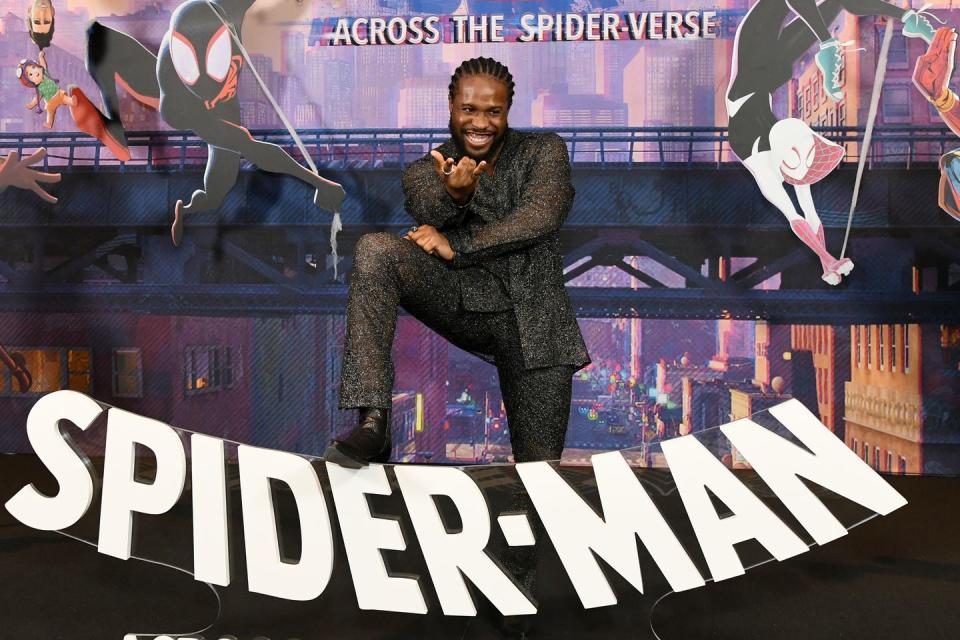 shameik moore gesturing while resting his right leg on top of a spiderman title logo