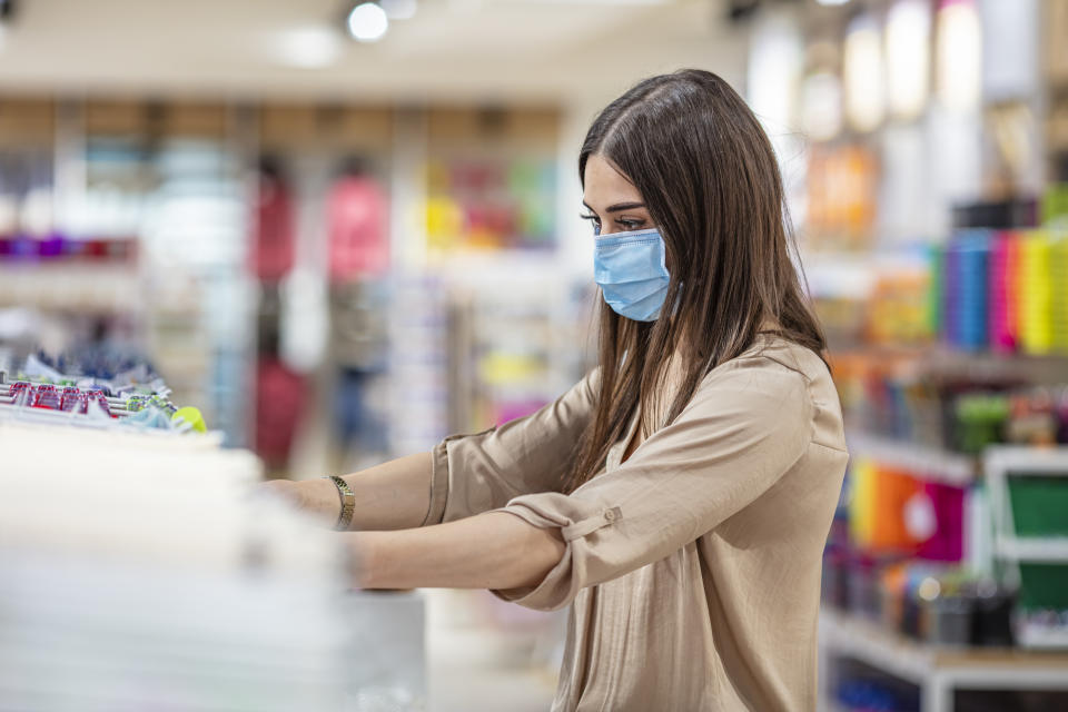 Woman wearing face mask push shopping cart in supermarket department store. Girl choosing, looking grocery things to buy at shelf during coronavirus crisis or covid19 outbreak.