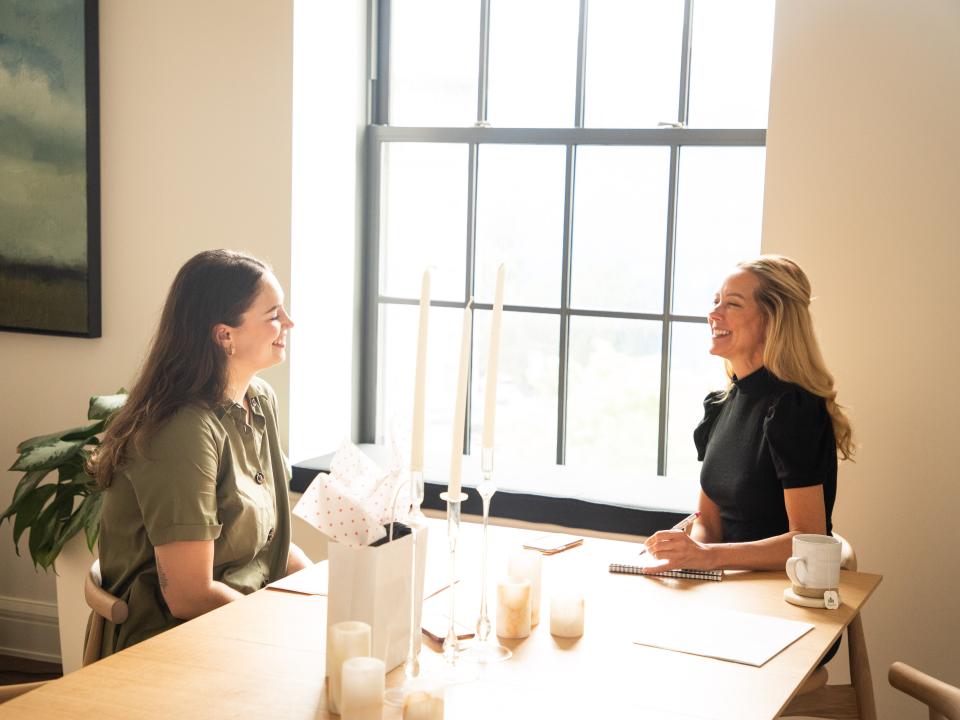 Two women sit at the end of a table and face each other while talking.