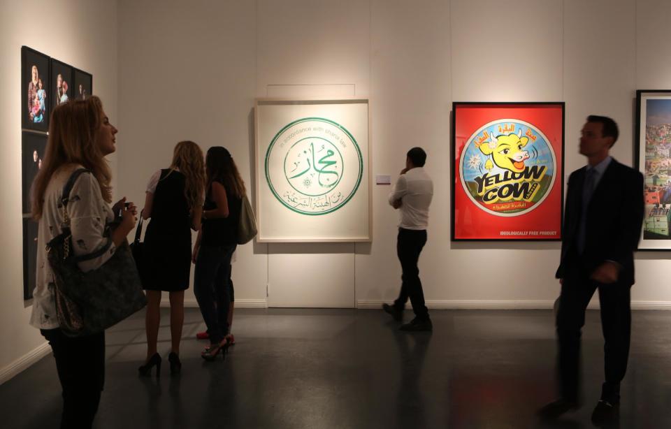 In this Monday, Sept. 16, 2013 photo, people view works of art, including a silkscreen print titled, "The Stam" by Saudi artist Abdulnasser Gharem, middle, and another titled, "Yellow Cow Cheese," by Saudi artist Ahmed Mater, right, during an auction at the Ayyam gallery, that moved from Damascus to Dubai in late 2011, in Dubai, United Arab Emirates. The auction Monday in Dubai's evolving art district served as a window into a small but forward-looking effort to save one niche of Syria's artistic community with no end in sight to the civil war that has already claimed more than 100,000 lives. (AP Photo/Kamran Jebreili)