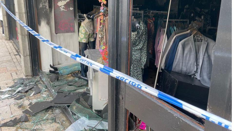 A smashed shop window and smoke damaged darkened walls with clothes on rails and a police cordon line 