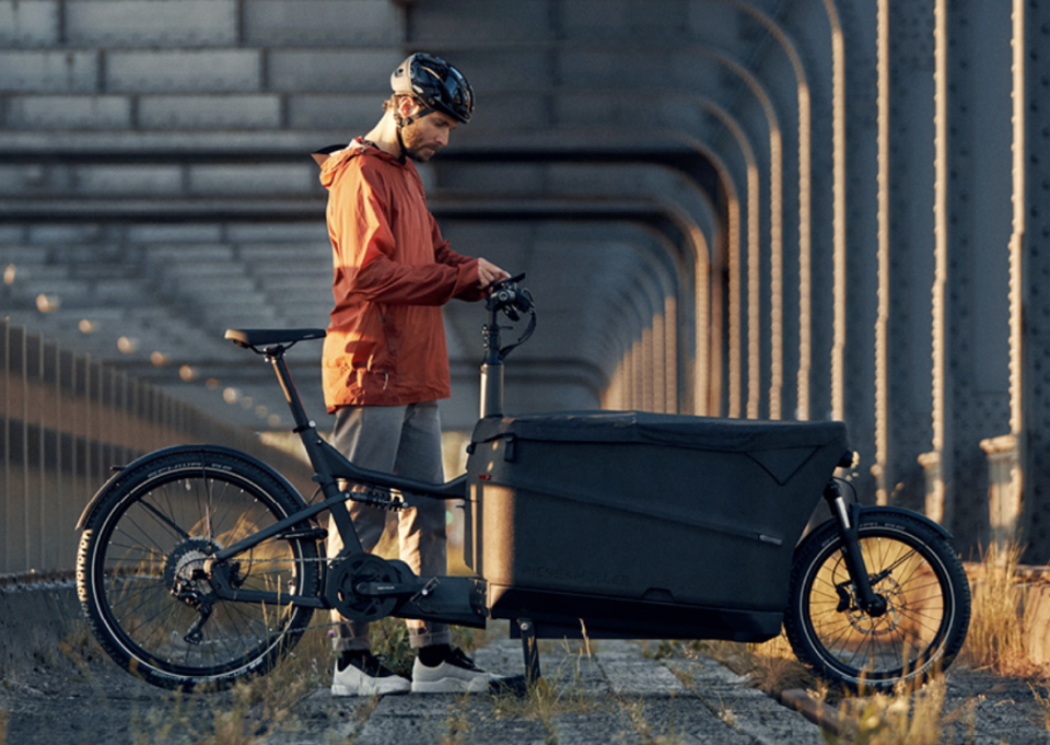 A man in an orange coat stands beside a Riese and Muller black Packster 70 in front of an industrial background