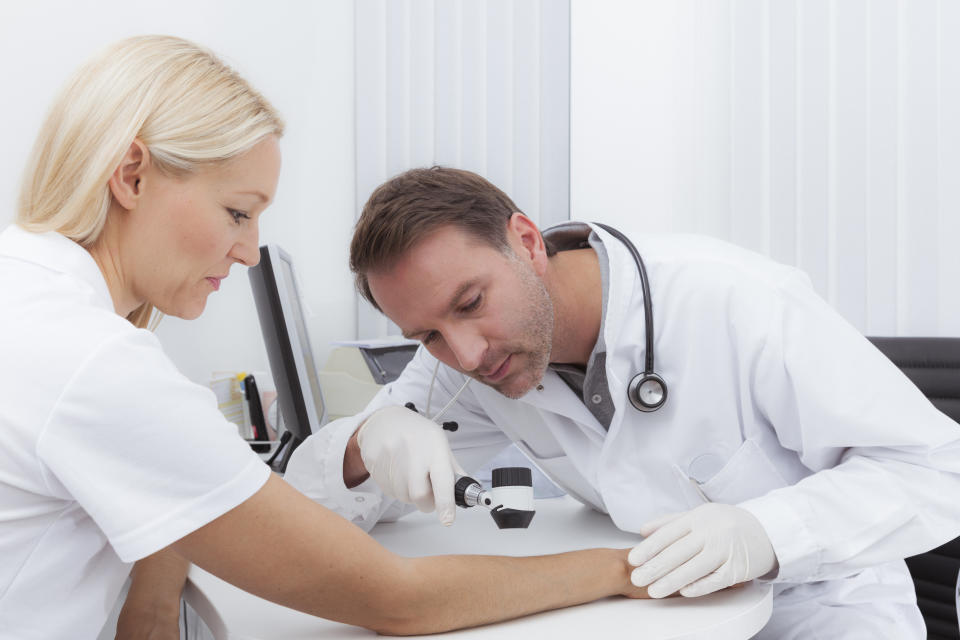 male dermatologist in lab coat looking at blonde woman's arm wearing white t-shirt, checking for skin cancer