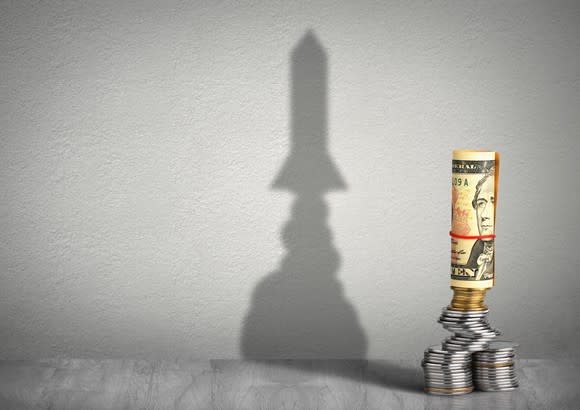 Coins and a rolled $10 bill are stacked to look like rocket ship with its shadow on the wall behind it.