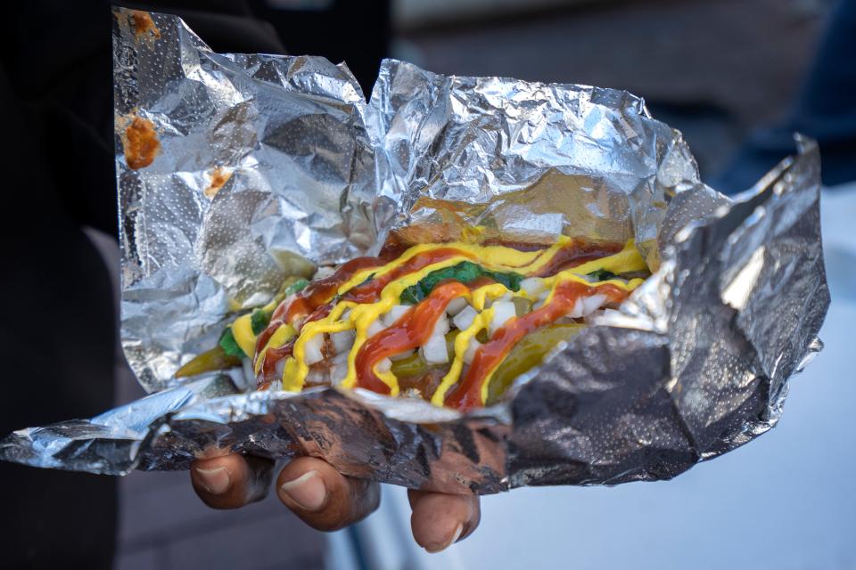 Torin Harris shows his beef polish hot dog with everyting that he got from Charles Smith’ Road Dogs cart on Monument Circle, Thursday, Nov. 2, 2023. Smith has been in this location since 2019 with his cart, Road Dogs, selling gourmet fusion street food. He loves the freedom and creativitiy of his small business. “A hot dog is a canvas. You can put whatever you want on it to make it special,” he says.