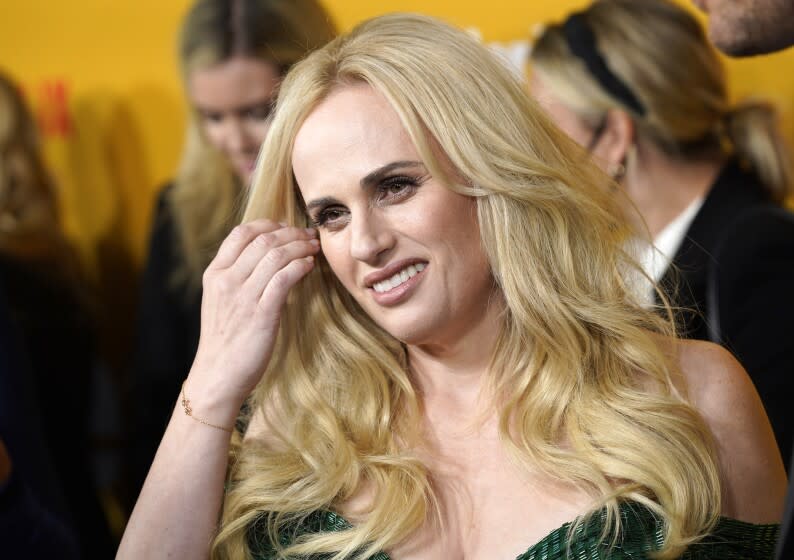 A woman pulls long hair back from one side of her face as she smiles gently for cameras at a movie premiere