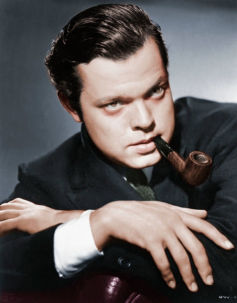 Orson Welles, in a suit with a pipe in his mouth, rests his arms on a surface and looks forward