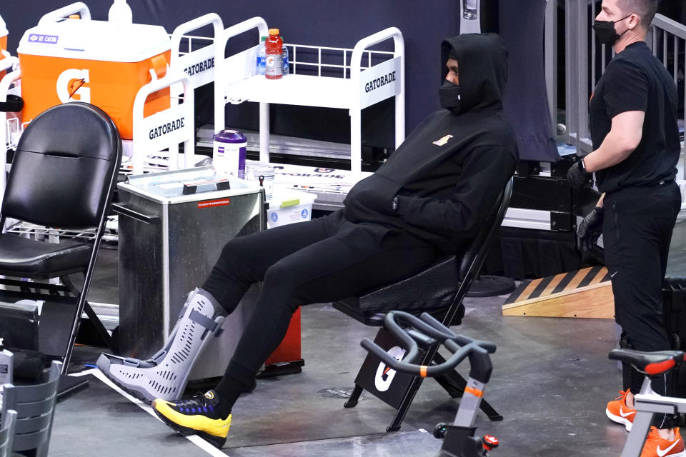 LeBron James sits in a chair near the bench area wearing black sweats with his hoodie pulled over his head and a black facemask with a walking boot on his right foot.