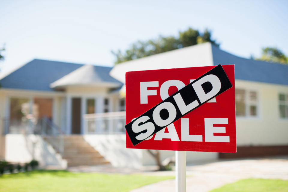 A "For Sale" sign in front of a house with a "Sold" banner across it, indicating the house has been purchased