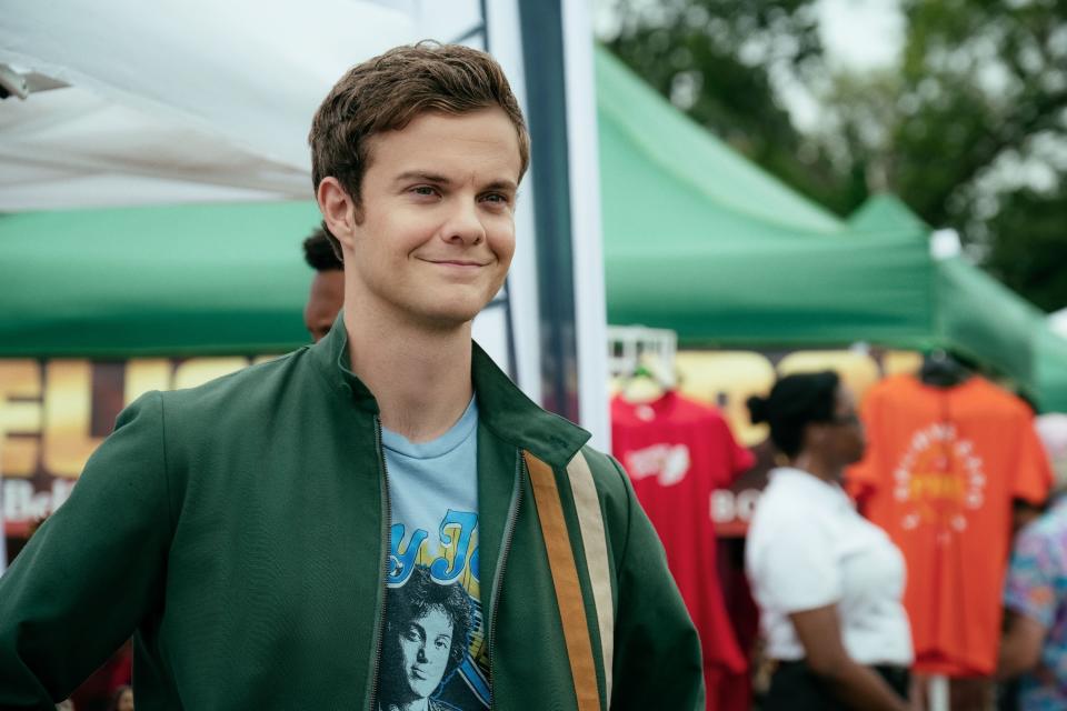Actor wearing a graphic tee and green jacket with a striped detail, standing at an outdoor event with tents and people in the background