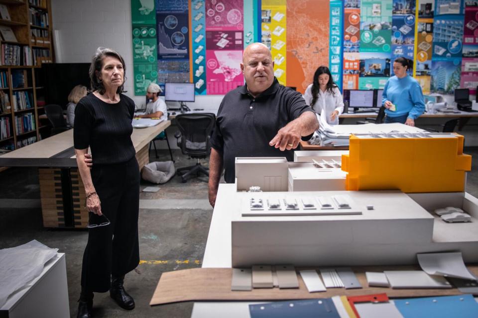 A woman in a black dress and a man with a shaved head stand behind a table filled with architectural models