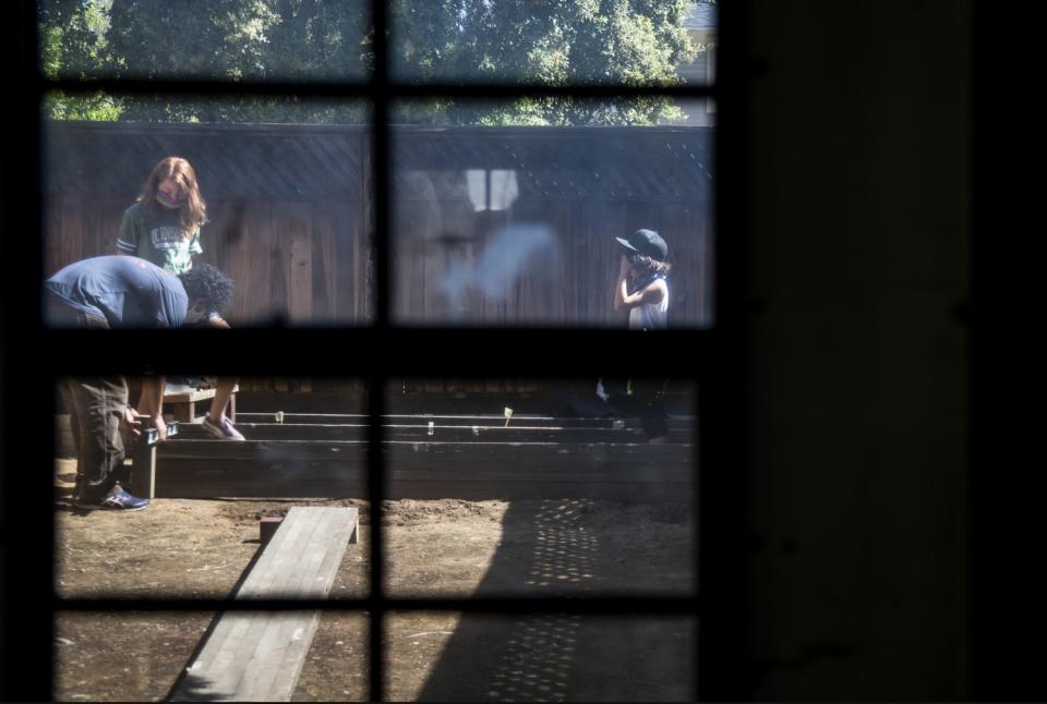 Seen through a shed window, Gabe Roth teaches Kat Bristow and Sylvester Roth how to build a raised bed in a gardening class.