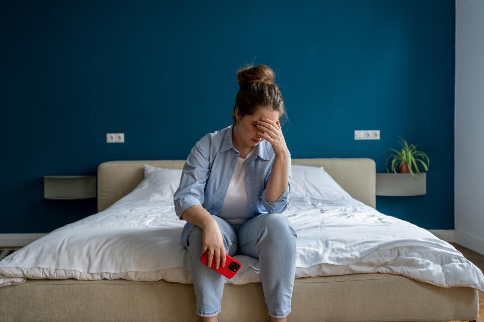 A woman sits on the edge of her bed and is holding her phone in one hand while the other is placed over her eyes as she leans her elbows on her knees