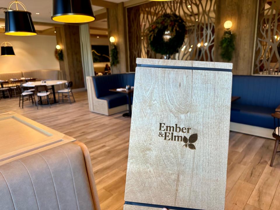 Wooden Ember & Elm menu being held in front of the restaurant's blue booths and light wood exterior