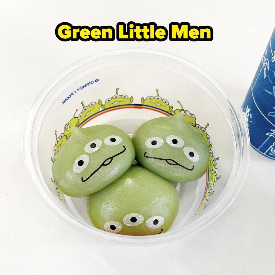 Three green mochi featuring the faces of Toy Story aliens with three eyes are in a plastic container with an alien-themed design