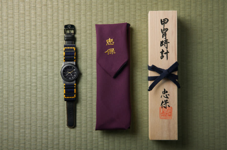 A bespoke watch made of traditional Japanese armor next to its cloth carrying pouch and wood case.