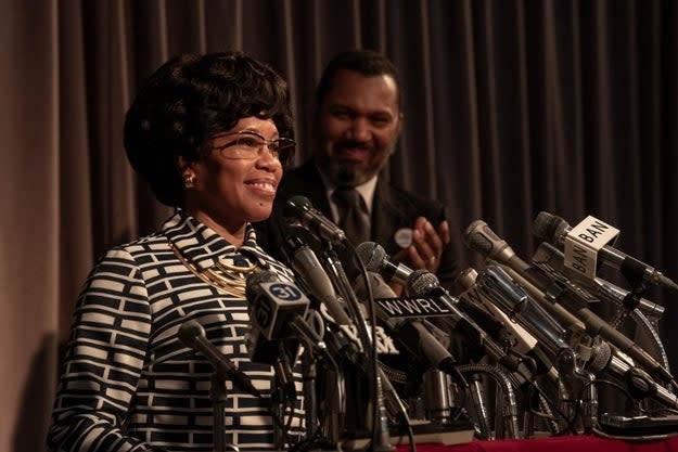 Woman in striped dress at podium, man smiling behind, both in formal attire, at apparent press event