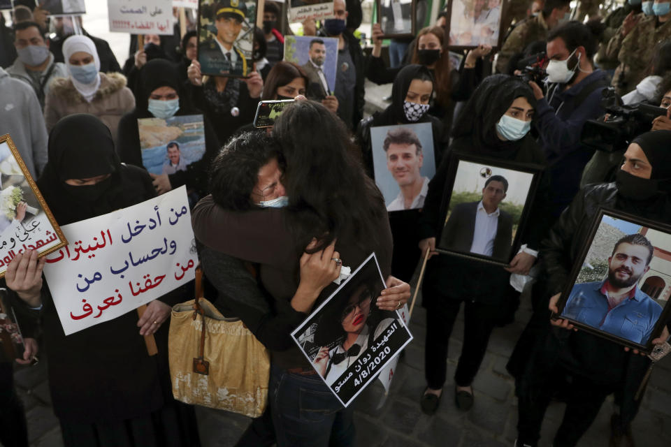 Two women embrace as relatives of victims of the Aug. 4, 2020 Beirut port explosion hold portraits of their loved ones who were killed during the explosion, during a vigil at the seaport main entrance, as they marked six months since the blast that killed more than 200 people and injured thousands, in Beirut, Lebanon, Thursday, Feb. 4, 2021. The Arabic words on poster read "Who brought nitrates and for who? We have a right to know." (AP Photo/Bilal Hussein)