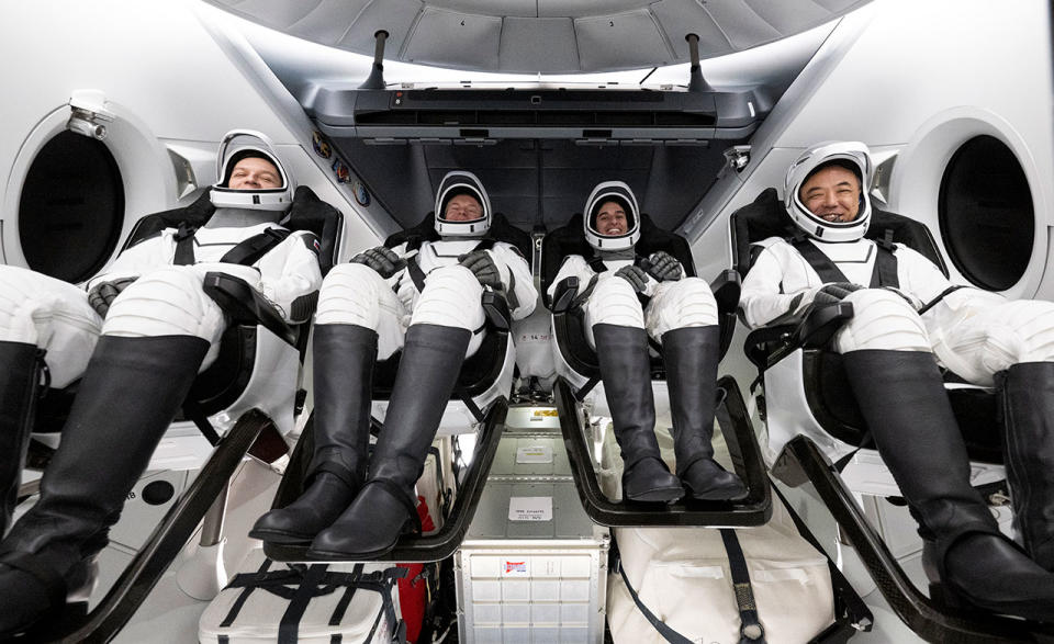 four people in black and white spacesuits sit inside a white space capsule