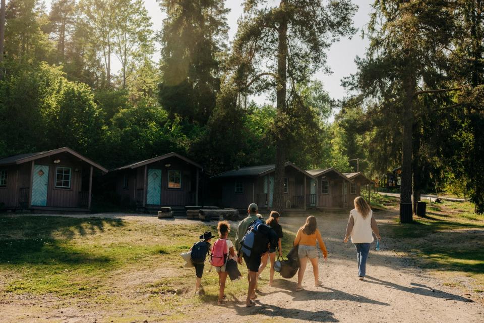 A family with backpacks and personal belongings approaches four cabins on a sunny day in the woods.