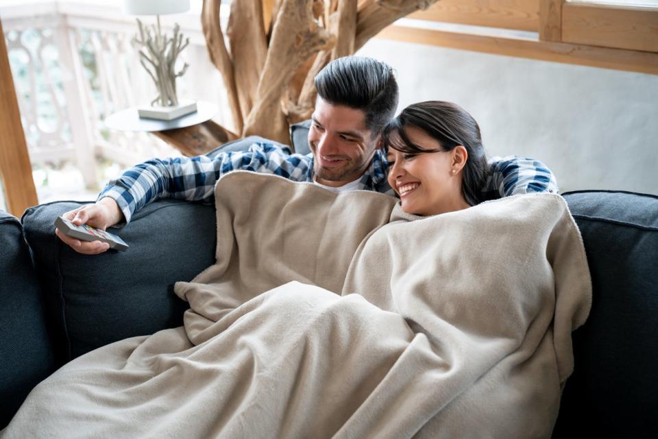 Man and woman on couch under a beige blanket, watching TV.