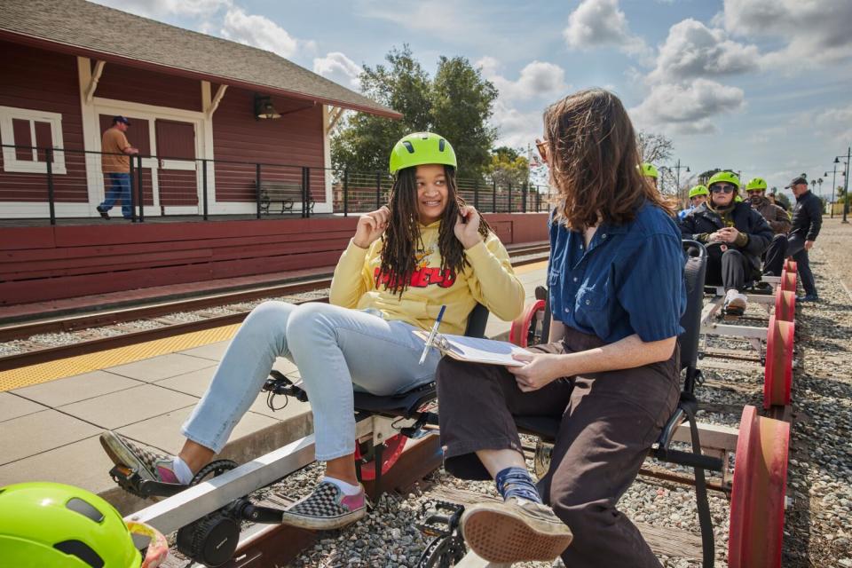 Two young people seated in a railbike, putting neon yellow helmets on and looking at each other.