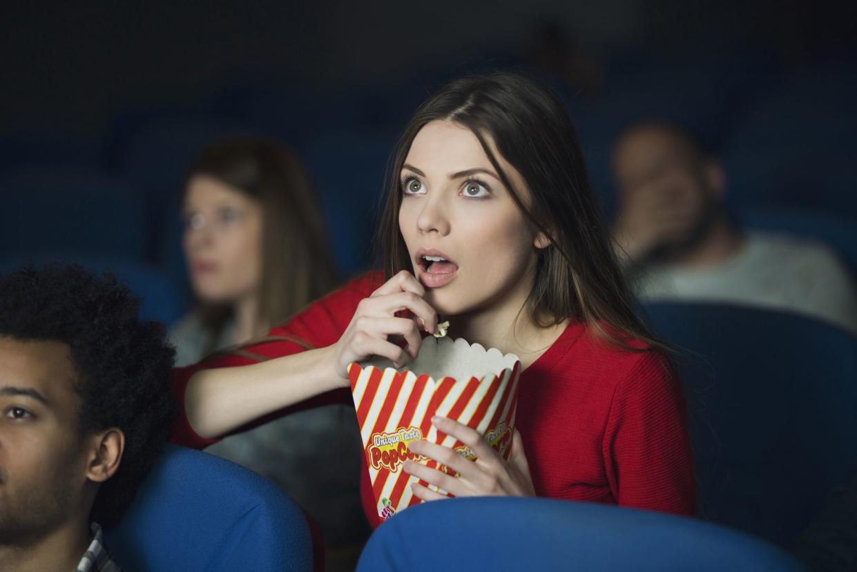 young woman eating popcorn on edge of seat in movie theatre with surprised expression