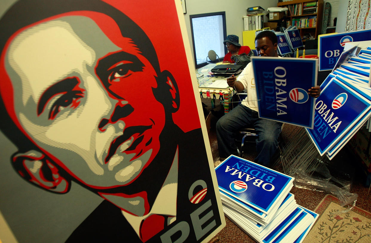 Surrounded by stacks of blue Obama Biden lawn signs and a large portrait of Obama subtitled Hope, Tony Smith works, with another African American volunteer in the background.