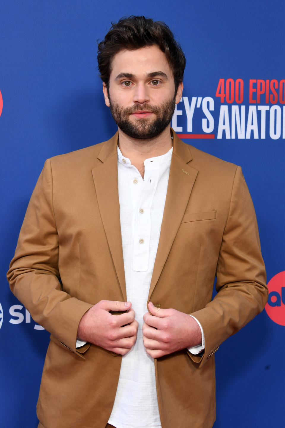 Jake Borelli posing at a Grey's Anatomy event, wearing a white linen shirt and a tan blazer