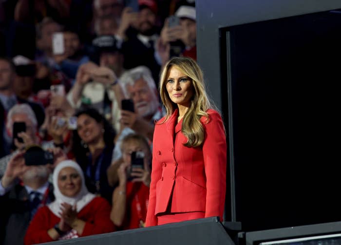 Melania Trump stands on stage wearing a tailored, double-breasted suit. She is in front of a crowd with many people taking photos