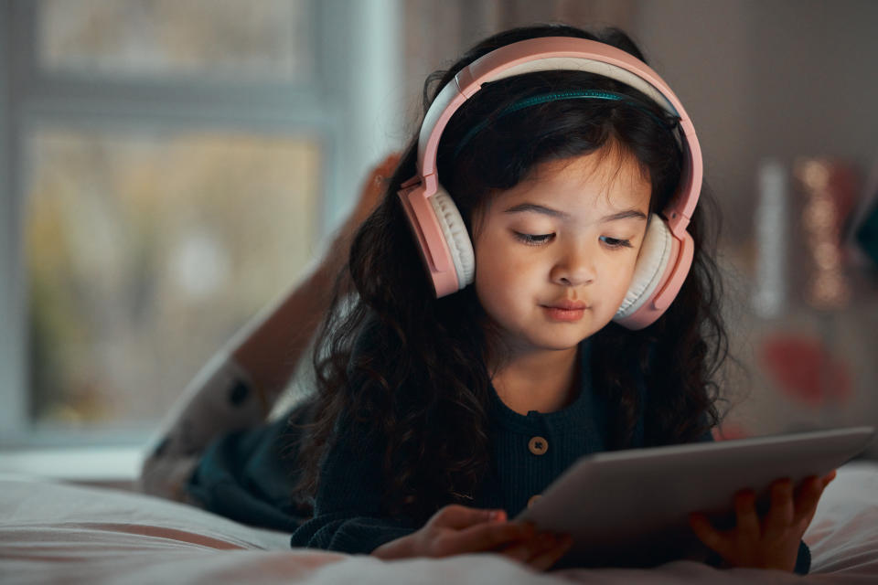 Child with long hair wearing headphones, lying on a bed while looking at a tablet
