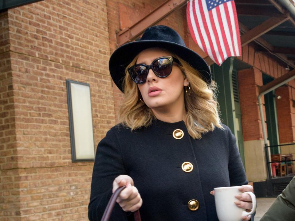 Adele with a hat and sunglasses on holding a mug of tea outside in front of a hanging American flag.