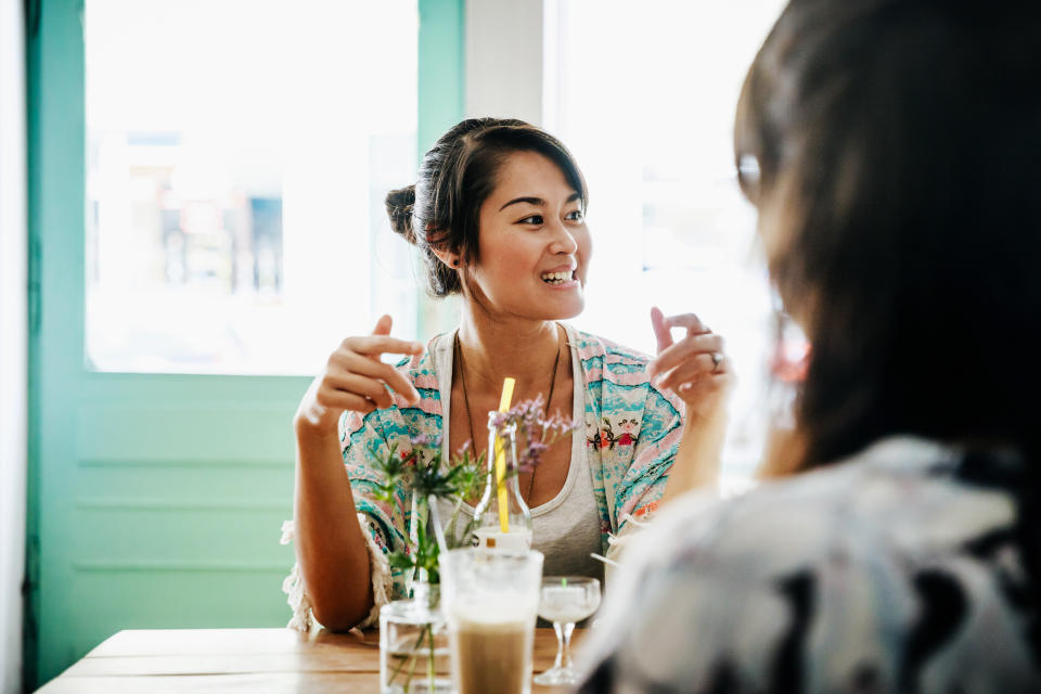 A young woman is sitting down for refreshments in a bright cafe, smiling and chatting with her friends.